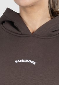 Zbliżenie osoby w ciemno-brązowej bluzie z kapturem z białym wyszywanym napisem "SMILODOX" na klatce piersiowej poniżej dekoltu.