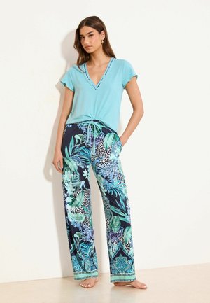 Frau steht barfuß, trägt ein hellblaues, kurzärmeliges V-Ausschnitt-Top und lockere dunkelblaue Hosen mit grünem und türkisfarbenem Blumen- und Tiermuster.