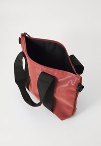Sac rouge, brillant, imperméable avec des poignées en nylon noir, fermeture éclair en haut, et un intérieur ouvert présentant un revêtement noir. Forme rectangulaire simple.