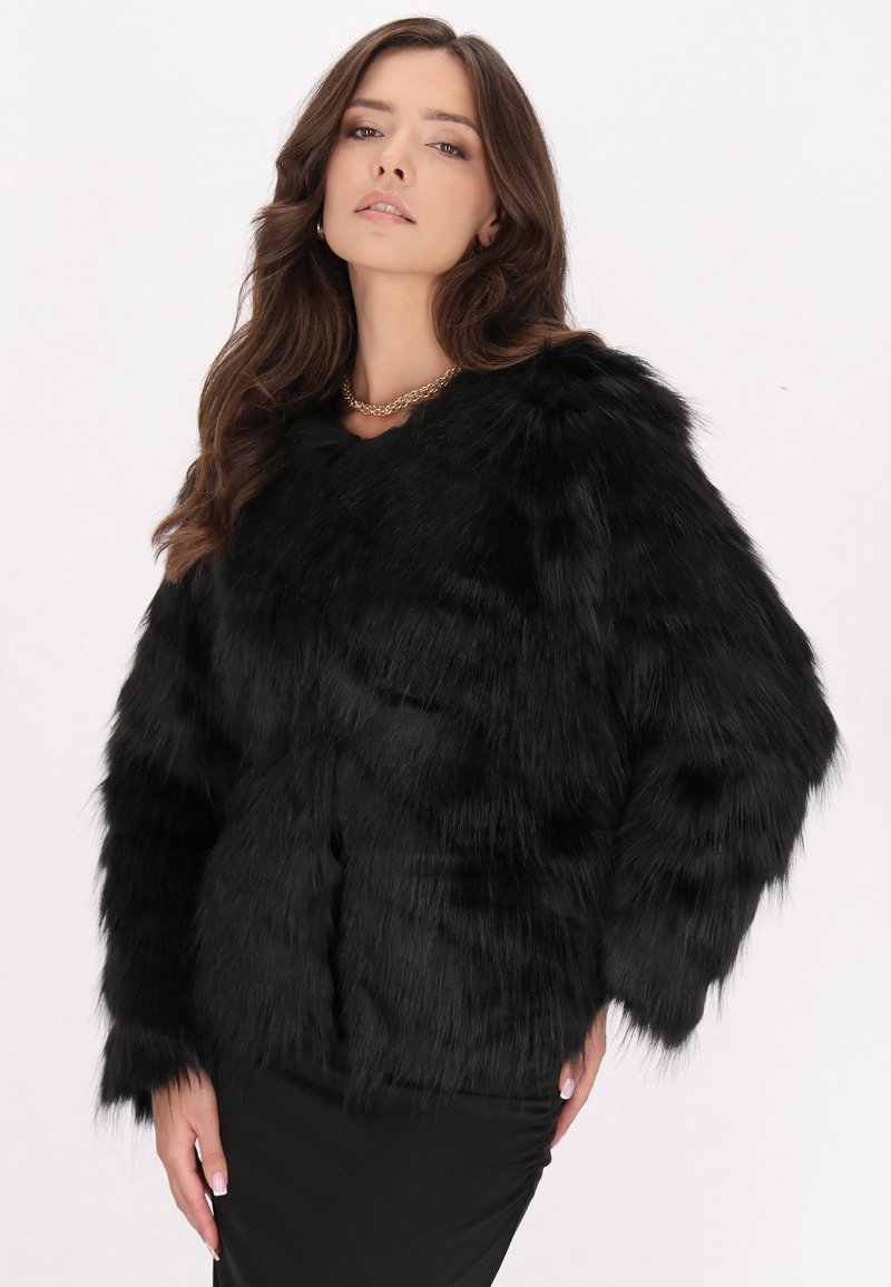Sort faux fur-jakke med en blød, tekstureret finish, der har brede ærmer og en rund halsudskæring, parret med en glat sort nederdel.