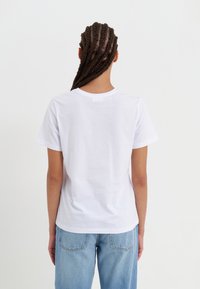 Camiseta de algodón blanca con cuello redondo y mangas cortas, con una textura suave. Llevada con jeans de mezclilla azul claro, mostrando la vista trasera.