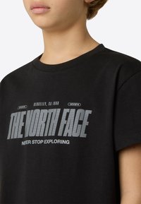 Fekete pamut póló, amelyen egy nagy szürke logó látható a "THE NORTH FACE" és a "NEVER STOP EXPLORING" feliratokkal. Rövid ujjú, kerek nyakkivágással.