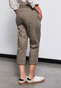 Khaki zkrácené kalhoty s elastickým pasem, bočními kapsami a detailními knoflíky na spodním lemu, doplněné o světlé tenisky.