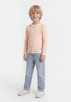 Usmievajúce sa blond dieťa stojace, oblečené v broskyňovo kockovanej dlhorukávovej košeli, svetlomodrých džínsach a bielych teniskách na bielom pozadí.
