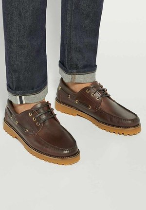 Brown leather shoes with a textured finish, featuring gold eyelets, brown laces, and a contrasting gum sole. Paired with dark denim jeans.