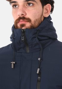 Giacca blu navy con zip, colletto alto, cordini regolabili e tasche con zip. La texture sembra liscia, con dettagli in metallo.