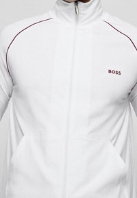 Giacca bianca con cerniera e collo alto, caratterizzata da accenti in bordeaux sulle spalle e logo BOSS sul petto. Realizzata in tessuto liscio.