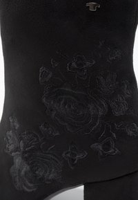Détail d'une botte en daim noir avec des motifs floraux brodés ton sur ton et un petit logo métallique près du bord supérieur.