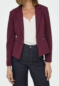 Blazer ajusté bordeaux avec une coupe cintrée, deux poches avant, revers à crans et manches longues, assorti à un haut blanc et un jean sombre.