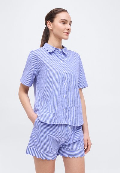 Γυναίκα που φοράει μπλε και λευκό πουκάμισο με κάθετες ρίγες και κοντά μανίκια κουμπωτό, μαζί με ταιριαστό σορτς με κυματιστό στρίφωμα, στεκόμενη με το ένα χέρι στην τσέπη.