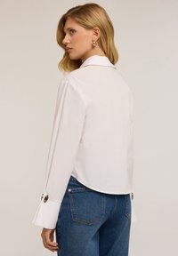 Camicia bianca a maniche lunghe con colletto classico e polsini svasati, con dettagli di bottoni argentati. Abbinata a jeans di denim blu.