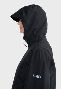 Veste noire imperméable avec capuche, dotée d'une fermeture éclair à l'avant et de boucles élastiques. Logo AIGLE imprimé en blanc sur la manche gauche. Texture lisse.