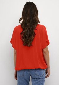 Rotes, locker sitzendes Oberteil mit kurzen Ärmeln, aus leichtem Stoff und mit glatter Textur. Kombiniert mit hochgeschnittenen blauen Jeans.