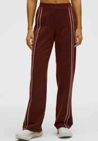 Brūnas sporta bikses ar sānu šuvēm kontrastējošā gaišā krāsā, ar maigu tekstūru un brīvu piegriezumu, kas ir piemērotas aktīvai lietošanai.