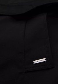 Czarna tkanina z przeszytym brzegiem i małą srebrną prostokątną metką z napisem "QUINTESSENCE" przymocowaną w pobliżu dolnego prawego rogu.