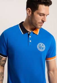 Niebieska koszulka polo z czarnym kołnierzykiem i pomarańczowymi akcentami. Posiada okrągłe logo na klatce piersiowej, zapięcie na trzy guziki oraz teksturowany materiał.