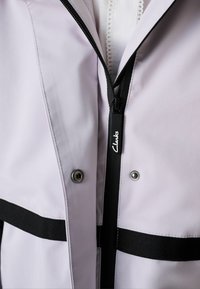 Svetlofialová nepremokavá bunda s čiernym zipsom a akcentmi, s kovovými patentkami a štruktúrovanou látkou s vodorovnými čiernymi pruhmi.