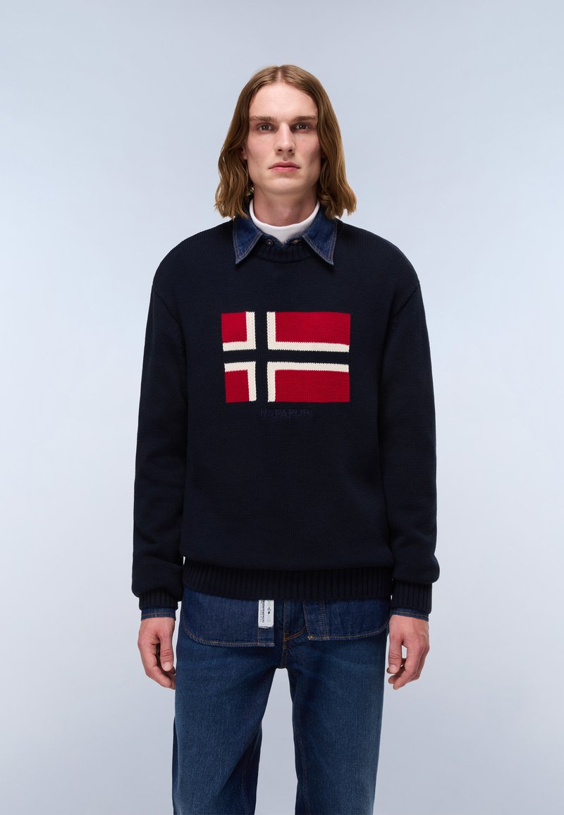 Joven con pelo hasta los hombros que lleva un suéter azul marino con diseño de cruces rojas, blancas y negras, camisa vaquera y vaqueros azules sobre un fondo liso.