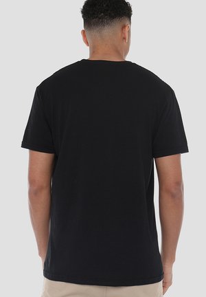 Man facing away wearing a plain black short-sleeve t-shirt and beige pants against a plain light gray background.