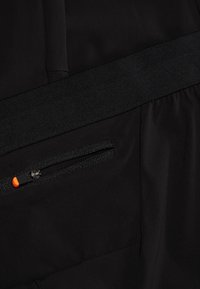 Zwarte stof met een gladde textuur, voorzien van een zwarte elastieken tailleband. Een ritszak heeft een zwarte rits en een oranje trekkoord.