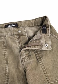 Gros plan de la ceinture d’un pantalon en velours côtelé kaki avec fermeture éclair partiellement ouverte, bouton métallique et étiquette noire de la marque Molo à l’intérieur.