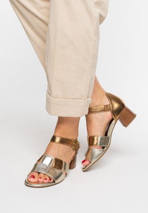 Feet wearing metallic gold and silver open-toe block heel sandals with ankle straps, paired with rolled-up beige corduroy pants.