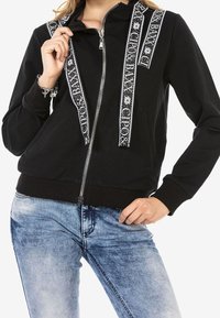 Black zip-up sweatshirt with contrasting white patterned straps; features a high collar and ribbed cuffs; paired with light blue jeans.