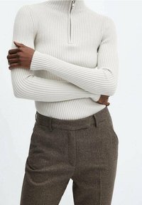 Pull à col roulé côtelé blanc avec une fermeture éclair moitié, associé à un pantalon marron texturé avec poches latérales et une silhouette ajustée.