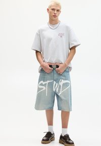 Grå oversized t-shirt med rødt logo, lyseblå denim shorts, brune lædersko og hvide sokker, med en fed hvid grafisk tekst.