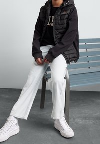 Czarna pikowana kamizelka z kapturem na czarnej koszulce, do tego jasnoniebieskie dżinsy i białe wysokie trampki, siedzący na niebieskiej ławce.