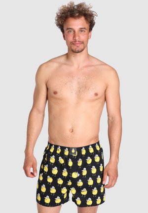 Schwarze Badekurze mit einem wiederholten Muster gelber Comicfiguren, elastischem Bund und Seitentaschen. Hergestellt aus leichtem Material.