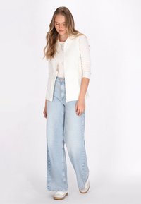 Weißer Zip-Westen über einem weißen Oberteil, kombiniert mit hellblauen, weiten Jeans. Das Model trägt weiße Schuhe mit beigen Akzenten. Geschmeidige Textur, lässiger Stil.