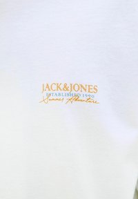 Tricou alb din bumbac, cu text brodat în portocaliu și albastru care spune "JACK & JONES Fondat 1990 Aventură de Vară." Textură moale.
