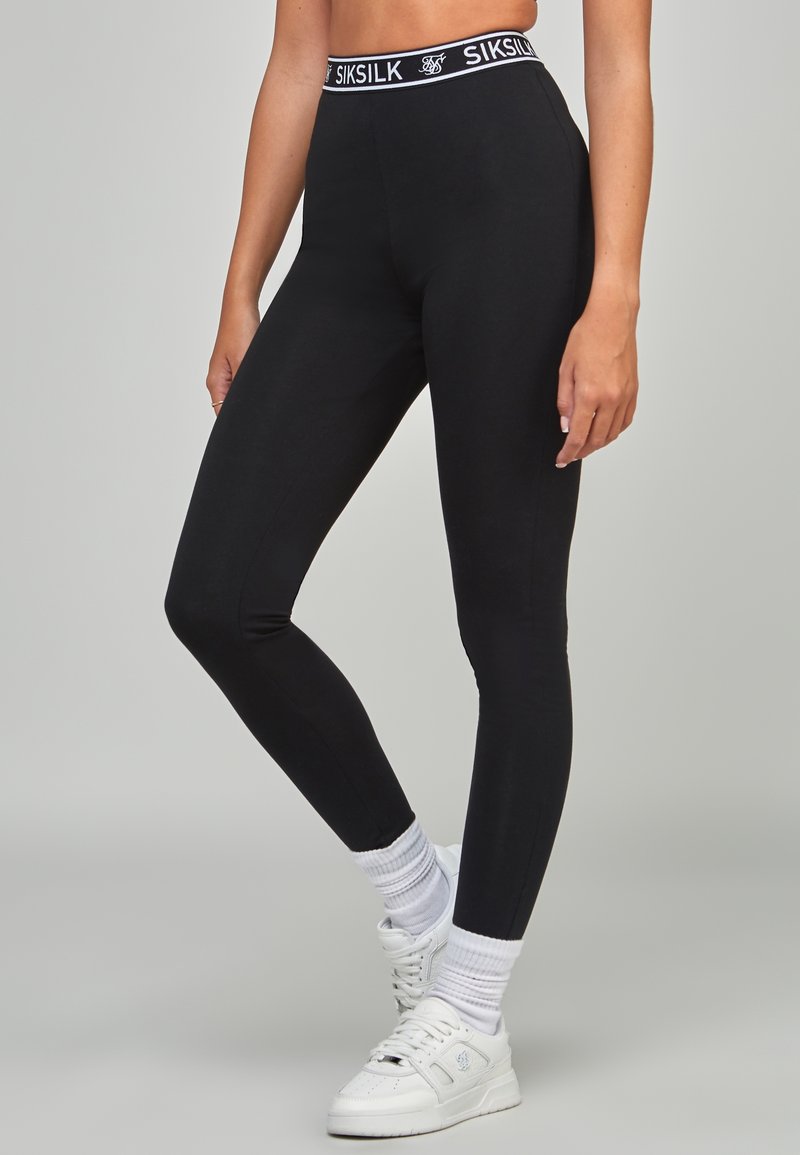 En person står i tajta svarta leggings med en märkesdesignad midjeband, tillsammans med vita sneakers och ankelstrumpor, mot en neutral bakgrund.