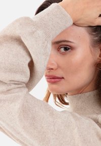 Beiger Rollkragenpullover mit gerippten Bündchen und Kragen. Weiche Textur, enganliegendes Design. Das Gesicht des Models ist teilweise neben dem Ärmel sichtbar.