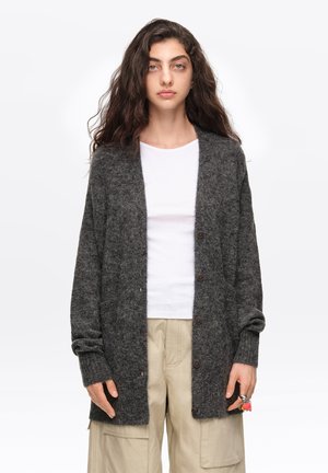 Joven con cabello largo y rizado lleva un cárdigan gris oscuro sobre una camisa blanca y pantalones cargo beige, de pie frente a un fondo blanco.