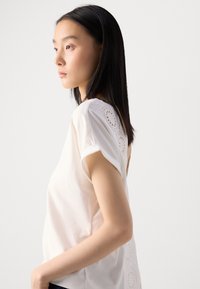 Chemisier blanc avec des manches courtes retroussées, des détails en broderie anglaise délicate et une texture lisse. Design simple et moderne.