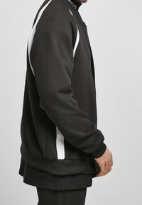 Chaqueta sudadera negra con franjas laterales blancas, puños ajustados y cuello redondo, fabricada con un tejido suave y texturizado. Diseño con cremallera.