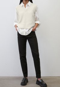 Helles, creamfarbenes Strickwesten über einem weißen Hemd mit Kragen, kombiniert mit dunklen, schmal geschnittenen Hosen und klobigen schwarzen Loafers. Einfaches und modernes Design.