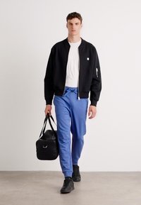 Zwarte bomberjack over een wit T-shirt, gecombineerd met blauwe joggingbroeken. De man draagt een zwarte sporttas en heeft zwarte sneakers aan.