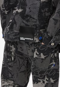 Μαύρο τζάκετ από τζιν με αφηρημένα γκρι μοτίβα, διαθέτοντας ένα μαύρο λογότυπο με την ένδειξη "KARL LAGERFELD JEANS" και μπλε λεπτομέρειες.