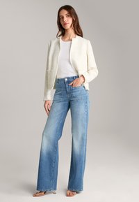Giacca bianca testurizzata, t-shirt bianca e jeans a gamba larga di colore azzurro chiaro. Dettagli notevoli includono ampie tasche sui jeans e una silhouette aderente.