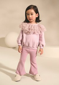 Sweat à lilas avec un col en tulle superposé, associé à un pantalon à pattes évasées côtelé assorti et des chaussons blancs en peluche, sur un fond neutre.