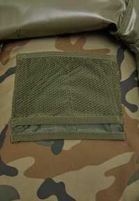 Olīvu zaļas kamuflāžas audums ar teksturētu acs kabatu. Kabatai ir pastiprinātas malas un gluda aizmugures materiāla.