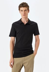 Polo negro de canalé con cuello, mangas cortas y corte slim, combinado con pantalones beiges. Diseño minimalista sin logotipos.