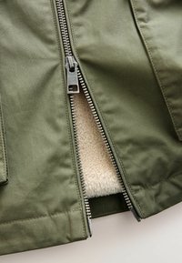 Olive green jacket with a silver metal zipper, featuring a soft beige fleece lining visible through the partially unzipped opening.
