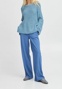 Femme portant un pull en tricot bleu, un pantalon bleu ample et des mocassins gris, debout les mains dans les poches sur un fond uni.