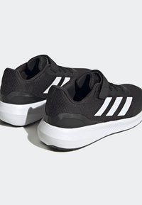 Baskets noires en tissu texturé, avec trois rayures blanches, un col rembourré et une semelle en caoutchouc blanche avec des détails.