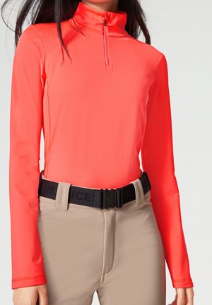 Vrouw droeg een fel koraalrode top met lange mouwen en een kwart rits, ingestopt in beige broek met een zwarte riem.