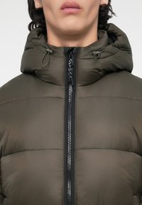 Chaqueta de plumas verde oliva con cuello alto y capucha ajustable con cordón. Cremallera negra central con detalles metálicos, textura suave.